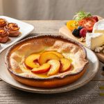 Styled Ontario brunch table with a peach Dutch baby, maple-glazed bacon roses, artisan cheese, fresh berries, and heirloom tomatoes on rustic plates under soft natural light.